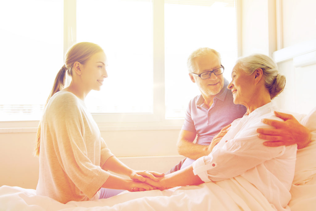 medicine, support, family health care and people concept - happy senior man and young woman visiting and cheering her grandmother lying in bed at hospital ward