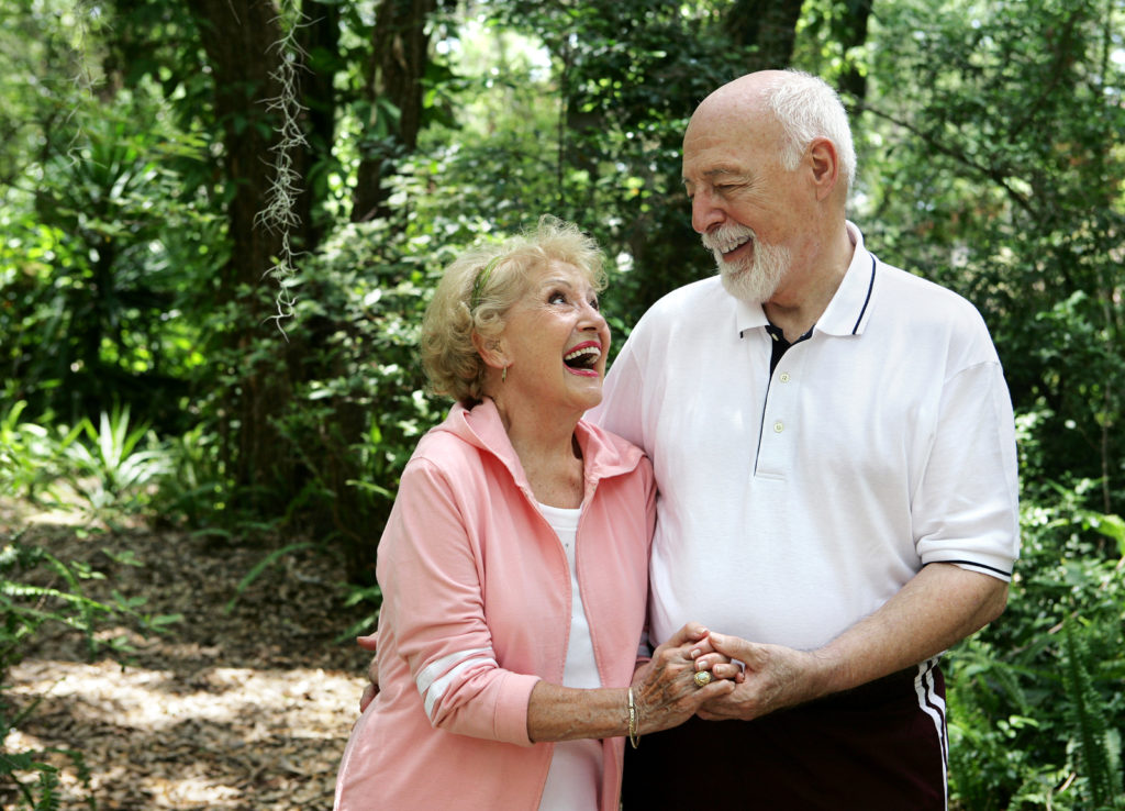 A happy active senior couple laughing together on a walk through the park. She's wearing a hearing aid. Plenty of copyspace.